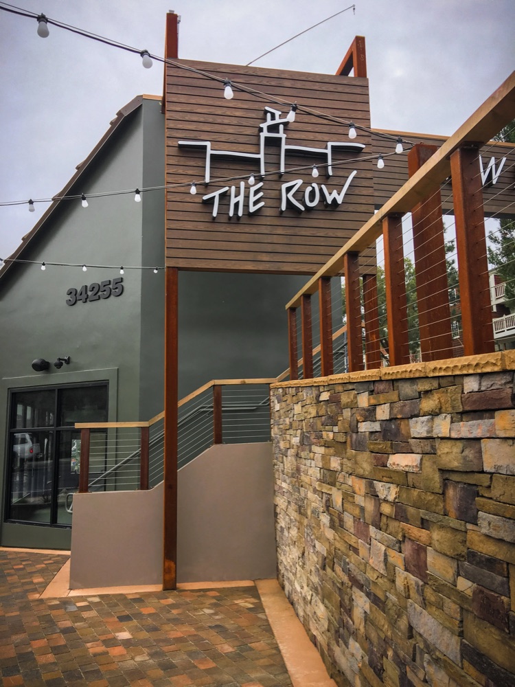 The Row blade sign mounted on a cedar gable, with string lights and a stacked-stone stair wall
